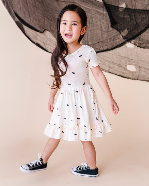 Young girl in a cream dress with halloween patterns and black shoes standing against a neutral background