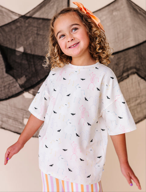 Young girl wearing a cream t-shirt with black bat patterns and striped pants, standing in front of Halloween-themed decorations.