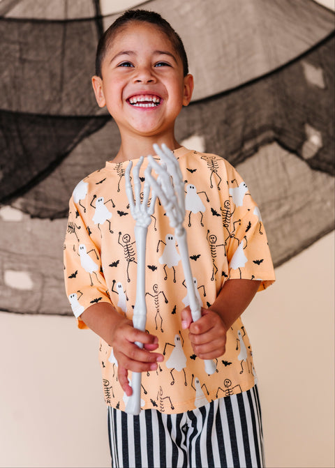 Child wearing a Halloween-themed shirt holding skeleton hands against a decorative backdrop.