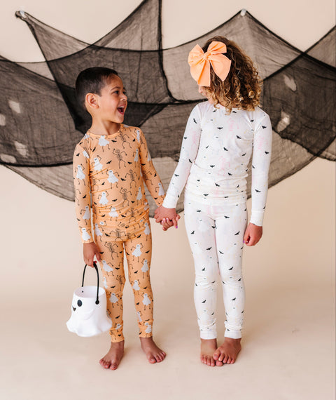 Two children in Halloween-themed pajamas standing against a beige background with decorative bats.