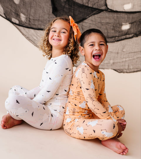 Two children sitting on a floor wearing pajamas with a halloween design.