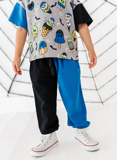 A child wearing a short-sleeve t-shirt with a colorful ice cream cone and ghost pattern on a gray base, paired with split black and bright blue pants and white sneakers, standing in front of a white web-like backdrop.