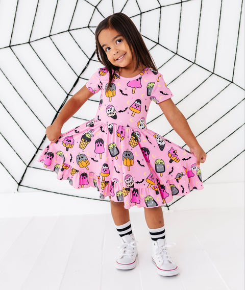 A young girl wearing a pink Halloween-themed dress with a spooky monster print, posing in front of a large black spiderweb prop on a white studio background.