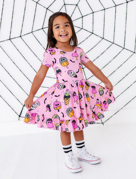 A young girl stands in a studio wearing a pink short-sleeve dress covered in a colorful Halloween print of pumpkins, ghosts, bats, and mummies. She is holding out the sides of the skirt to show its volume, with white socks and white sneakers, against a white background with a black spiderweb graphic.