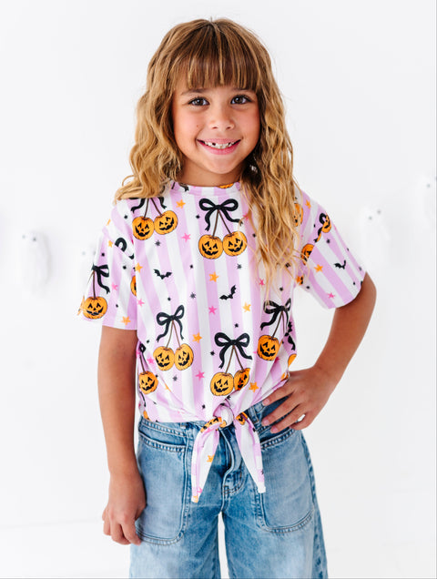 A young girl with blonde hair smiling while wearing a pink and white striped short-sleeve t-shirt with a Halloween pumpkin print, paired with light blue jeans, against a white wall.