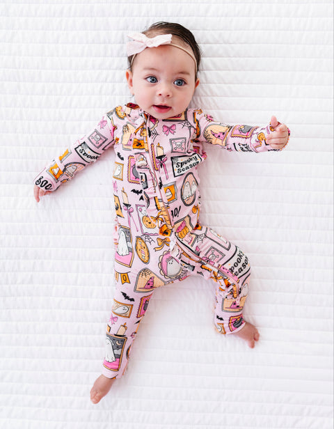 A baby lying on a white quilted blanket, wearing a pink long-sleeve footless onesie with a colorful illustrated print and a matching pink bow headband.