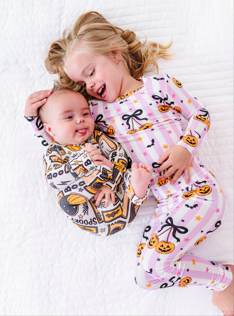 Two children lying on a white quilted bed wearing matching Halloween-themed long-sleeve pajamas. The older child has blonde hair and wears a light pink pajama set with black bows, orange pumpkins, and small stars.