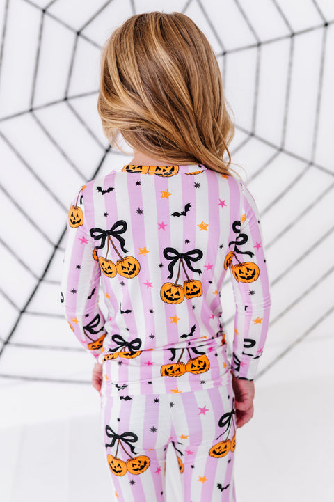 A child viewed from behind wearing a pink and white striped pajama set with a Halloween print of pumpkins, black bows, bats, and stars, set against a white background with a black spiderweb graphic.