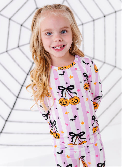 A young girl with blonde hair wearing a pink and white striped Halloween pajama set with jack-o'-lanterns, black bows, and bats, standing in front of a white background with a black spiderweb graphic.